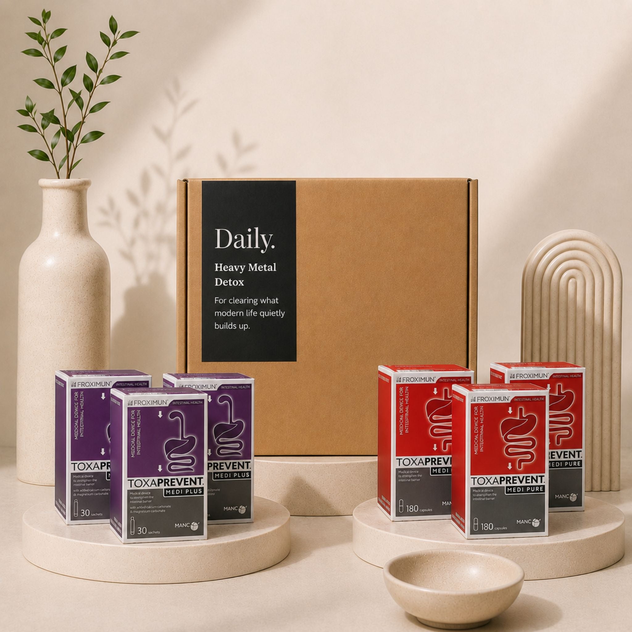 A detox supplement set displayed in a styled studio setup. The set includes a large brown cardboard box labeled 'Daily Heavy Metal Detox' in the background, with multiple individual product boxes in purple and red in the foreground on round pedestals.