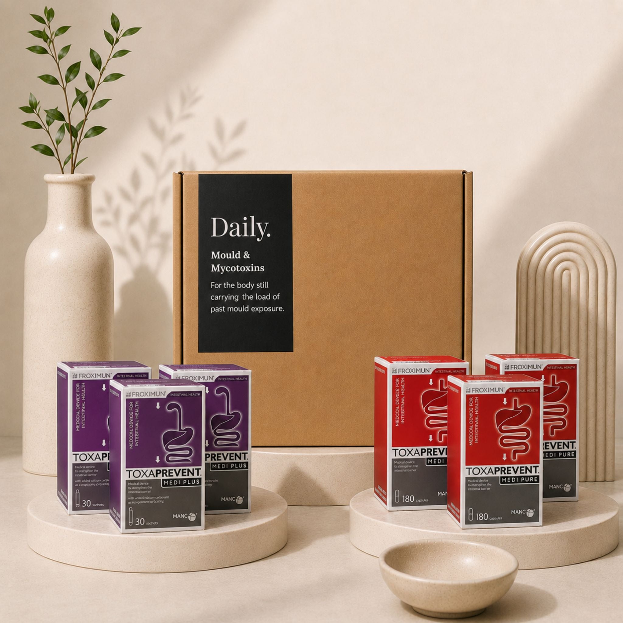 A Daily Tox Prevent subscription box set featuring a large brown cardboard box in the center, flanked by purple and red individual boxes of Tox Prevent and Toxaprevent on white pedestals, styled with a ceramic vase and a small bowl.